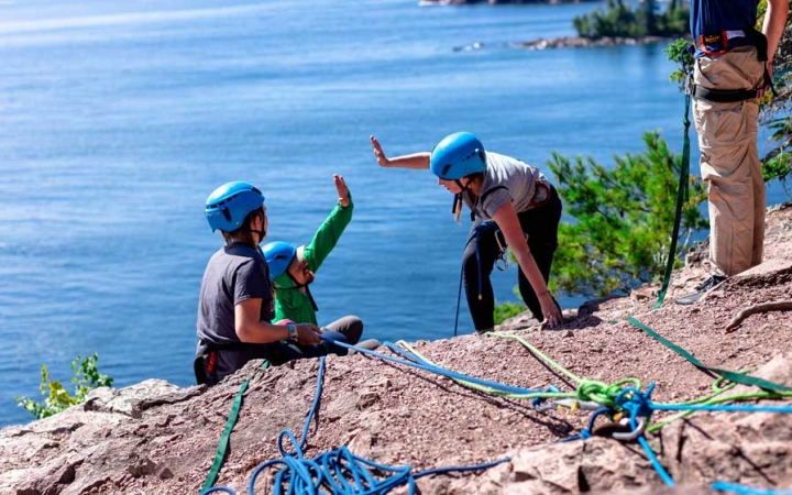Two students wearing safety gear and secured by ropes high five on the edge of a cliff, high above a large blue lake. 